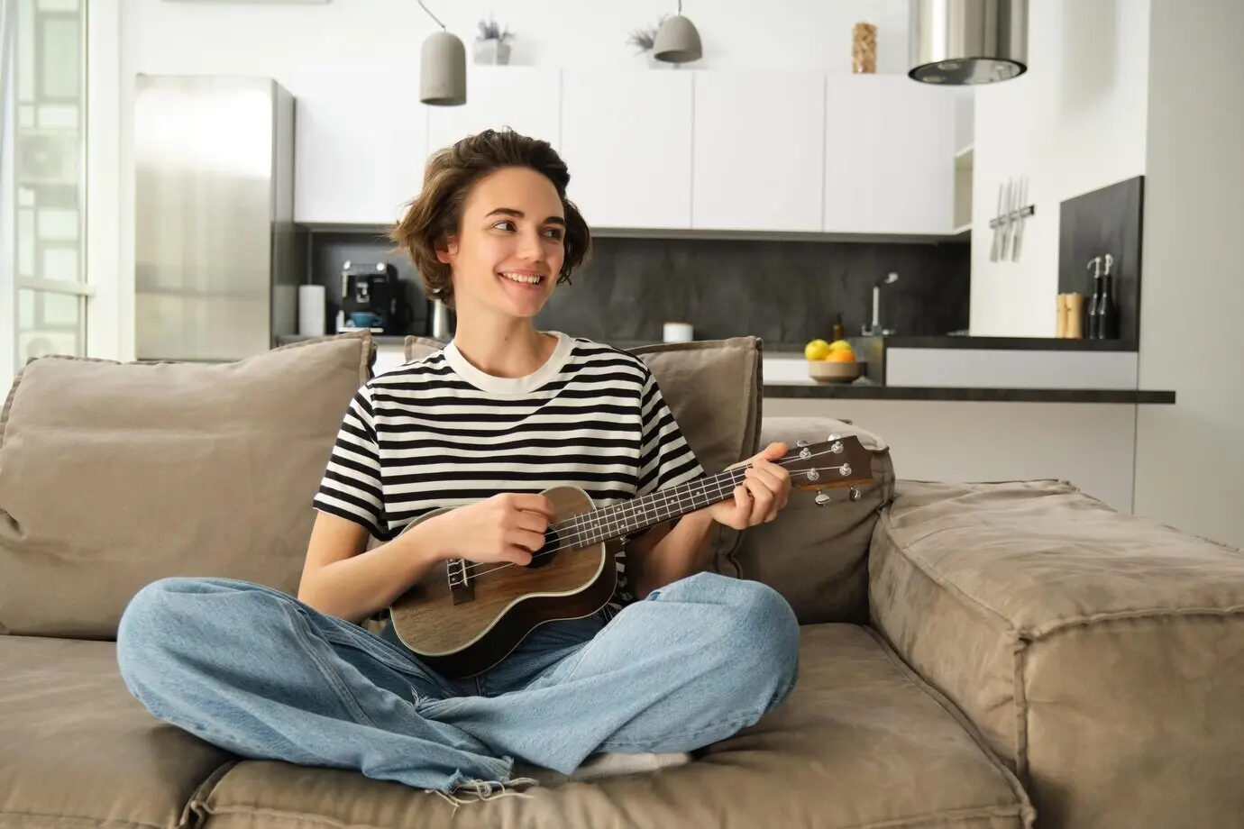Porträt einer niedlichen jungen Frau, die auf einem Sofa sitzt und lernt, Ukulele zu spielen, während sie ihr Musikinstrument hält.